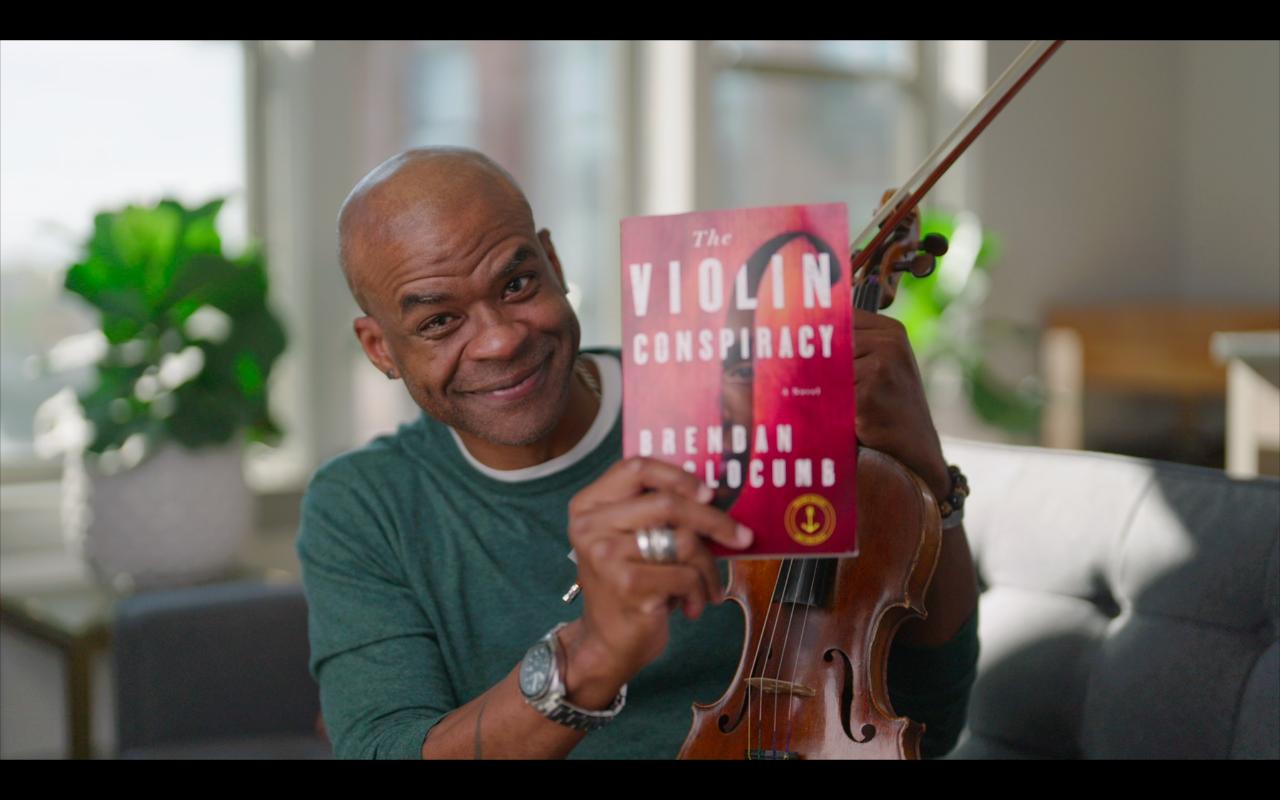 A picture of the author, a man in a teal shirt with a watch on and a gray tshirt underneath his teal shirt. He is holding a copy of the Violin Conspirary (with a red cover) and a violin. There is a room in the background blurred out with plants next to a window.